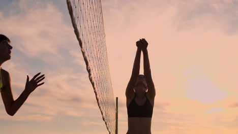 Cámara-Lenta,-ángulo-Bajo,-Primer-Plano,-Destello-De-Lente:-Jóvenes-Jugadoras-De-Voleibol-Pasan-Y-Rematan-La-Pelota-Sobre-La-Red-En-Una-Soleada-Tarde-De-Verano.-Chicas-Caucásicas-En-Forma-Jugando-Voleibol-De-Playa-Al-Atardecer.